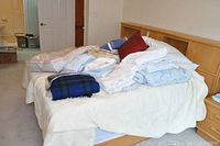 Bed with ivory bedspread with piled linens including quilts, comforters and sheets in assorted colors and patterns