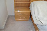 Photo of one wooden bedside table beside a bed, showing two drawers with wooden handles and an open shelf above the drawers.