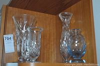 Five assorted clear glass vases placed on wooden shelf, showing a variety of shapes, sizes, and detail patterns. One tall vase has visible crack near base.