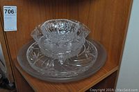 Three-piece set of clear glassware including a large plate, a medium bowl with triangular rim handles, and a small three-legged bowl with cut glass details on a wooden shelf.