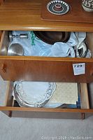 Wooden drawer open showing assorted silverplate serving items wrapped and stacked inside including trays, plates, candlesticks, bowls.