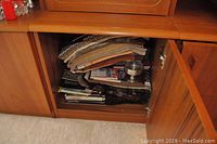 Cabinet shelf view showing stacked folded tablecloths on top, magazines underneath, a small fur-like rug, and a metal candle holder.