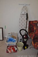 Ironing board with floral cover, upright Shark Euro Pro X vacuum cleaner, plastic laundry basket with storage bags and sewing patterns, beige waste bin, and folded fabric in red and white.