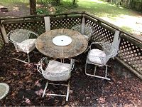 Wide view of outdoor metal mesh table surrounded by four white metal mesh chairs with padded white cushions on seat area, placed on leaf-covered ground with lattice fence background, showing outdoor sheeting and rust spots on metal surfaces.