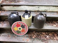 Photo showing three ceramic jugs and one round PA Dutch sign with leaf patterns on wooden steps surrounded by dry leaves.