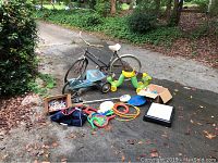 Wide view of lot items arranged on driveway showing bike, tricycle, inch worm toy, frisbees, hoops, metal detector, chalk box, and exercise step