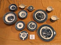 Top-down view of vintage Currier and Ives 40-piece dinnerware set with plates, cups, creamer, sugar bowl, and ashtrays displayed on yellow cloth