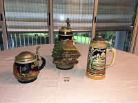 Three German beer steins displayed on a white cloth-covered table near window with outdoor view.