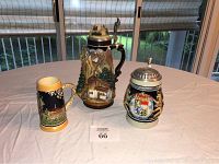 Front view of three German beer steins and mug on a table in front of window showing detailed designs and size differences.