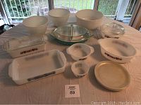 Front view of collection of vintage Pyrex and Fire King white opaque glass cookware and dinnerware on a beige tablecloth, including mixing bowls, casserole dishes, plates, small bowls, and a clear divided glass dish.