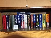 Bookshelf holding 18 Tom Clancy books arranged side by side with visible titles on spines.