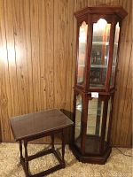 Front view of both curio cabinet with glass shelving and wooden end table, shown against wood paneled wall.