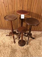 Four wooden pedestal stands arranged against a wood-paneled wall on a beige carpet. Three have tripod legs, one has a round solid base. Heights vary from 18 to 27.5 inches.