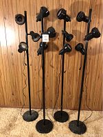 Front view of four black floor standing light fixtures with three adjustable lamp heads each, displayed against a wood-paneled wall.