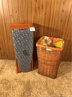 Wooden folding ironing board cabinet in closed position next to woven laundry hamper with fabric liner.
