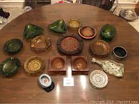 Entire assortment of 16 ashtrays showing variation in colors, shapes and materials on a wood table.