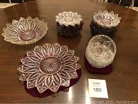 Entire glassware lot arranged on wooden table consisting of one large 11.5 inch plate, one large 11.5 inch bowl, twelve smaller 6 inch plates and thirteen small glass bowls. All pieces are clear with floral/starburst pattern cut designs.