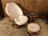 Photo showing the rattan swivel rocking chair with cream cushions, round ottoman, and glass top side table set in front of wood panel walls on beige carpet.