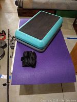 Blue and black 2-tier step platform on purple foam mat with tear; black ankle weights on mat; assorted weight plates and bars around.