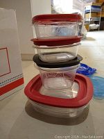 Stack of five small clear plastic food storage containers with red lids on a concrete floor beside a cardboard box