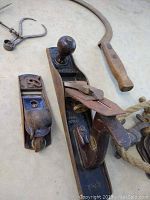 Close-up image of a vintage Stanley router plane with leather sanding strap alongside another antique wooden block plane, and part of a wooden-handled sickle visible in the background.