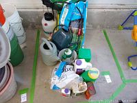 Image showing various gardening supplies including watering cans, bottles of liquids, fertilizer bag, and garden sprayers.