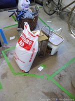 Photo of two bird feeders (wooden and metal) plus a large bag of wild bird seed labeled 'Wild Bird Feed'. Items placed on a concrete floor with bicycles in background.
