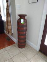 Photo shows tall brown floor vase with horizontal darker stripes placed on tile floor next to a wall and window.