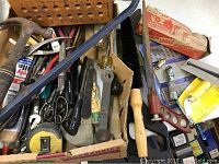 Assorted hand tools in a box including hammer, hacksaw frame, tape measure, pliers, scissors, and brushes.