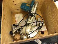 Overview of Makita compound mitre saw inside a wooden crate, showing saw, cord, and miscellaneous items.