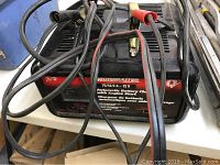 Motomaster automatic battery charger with cables and clamps shown from front angle highlighting product label and attached cables.