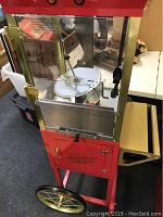 Front side showing glass compartment with popcorn stirring mechanism inside and red metal frame with gold handle and large wheels.