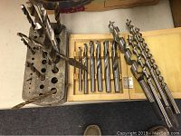 Overall view of various drill bits including auger and forstner bits shown on table with wooden case and metal holder