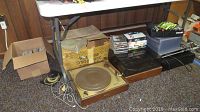 Photo showing two vintage Weathers phonographs on floor under table, one with wood base and beige platter, alongside cardboard boxes with CDs and tapes