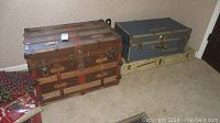 Photo of the large wooden vintage steamer trunk next to the smaller blue camp trunk and the metal box on floor.