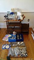 Wood tea cart with various kitchen and serving items displayed on and around it, including flatware, containers, and cloths.