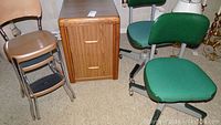 Two wheeled industrial chairs with green cloth seats and vinyl backs next to a woodgrain file cabinet and tan metal stool.