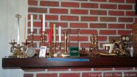 Wide view of assorted brass candle holders with candles, shown against brick wall background.
