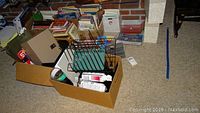 Photo showing a cardboard box with wire file organizer, spray bottles, and scattered office supplies on the floor with other boxes and items in the background.
