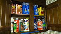 Photo showing bottles of cleaning liquids, sprays, and wood treatment products arranged on a wooden shelf.