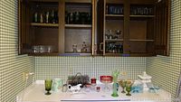Wide view showing various glass items on countertop and displayed in open cabinet. Items include colored goblets, bowls, and decorative glass pieces.