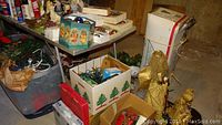 Overview of multiple boxes and bins containing Christmas decorations including angels, ornaments, and other holiday items on and around a table in a basement setting.