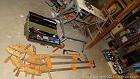 Photo showing two large wooden Jorgensen bar clamps with orange locks, a black toolbox with assorted tools, and other workshop equipment around.