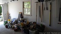 Wide shot showing garden tools leaning against wall, wheelbarrow, gardening products and other items organized on ground in garage.
