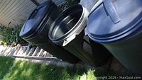 Three black plastic outdoor trash cans with lids, showing dirt and soiling.