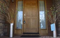 Wide angle photo showing two metal textured aluminum planters, each containing artificial mixed foliage with green leaves and dried sticks, positioned on the porch at either side of an external door.