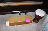 Wooden tongue drum alongside small hand drum, showing relative sizes and placement on carpeted floor under a piano bench
