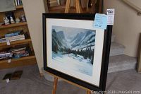 Framed watercolor painting showing mountain landscape with lake and snow, displayed on an easel.