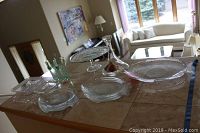 16-piece pressed glass lot displayed on a counter including fish plates, cake stand, glasses, decanter, and other plates