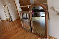 Two mirrors leaning against a wall on wooden flooring; one silver-framed rectangular mirror and one larger wood-framed mirror with an oval top